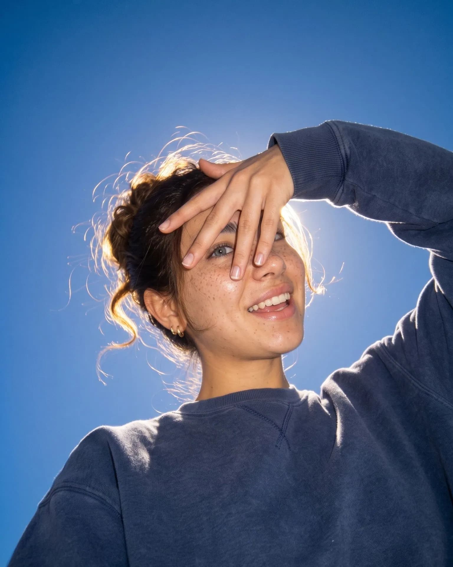 Freckled woman laughing in a navy sweatshirt against a deep cobalt sky, one hand spread over her forehead and one eye