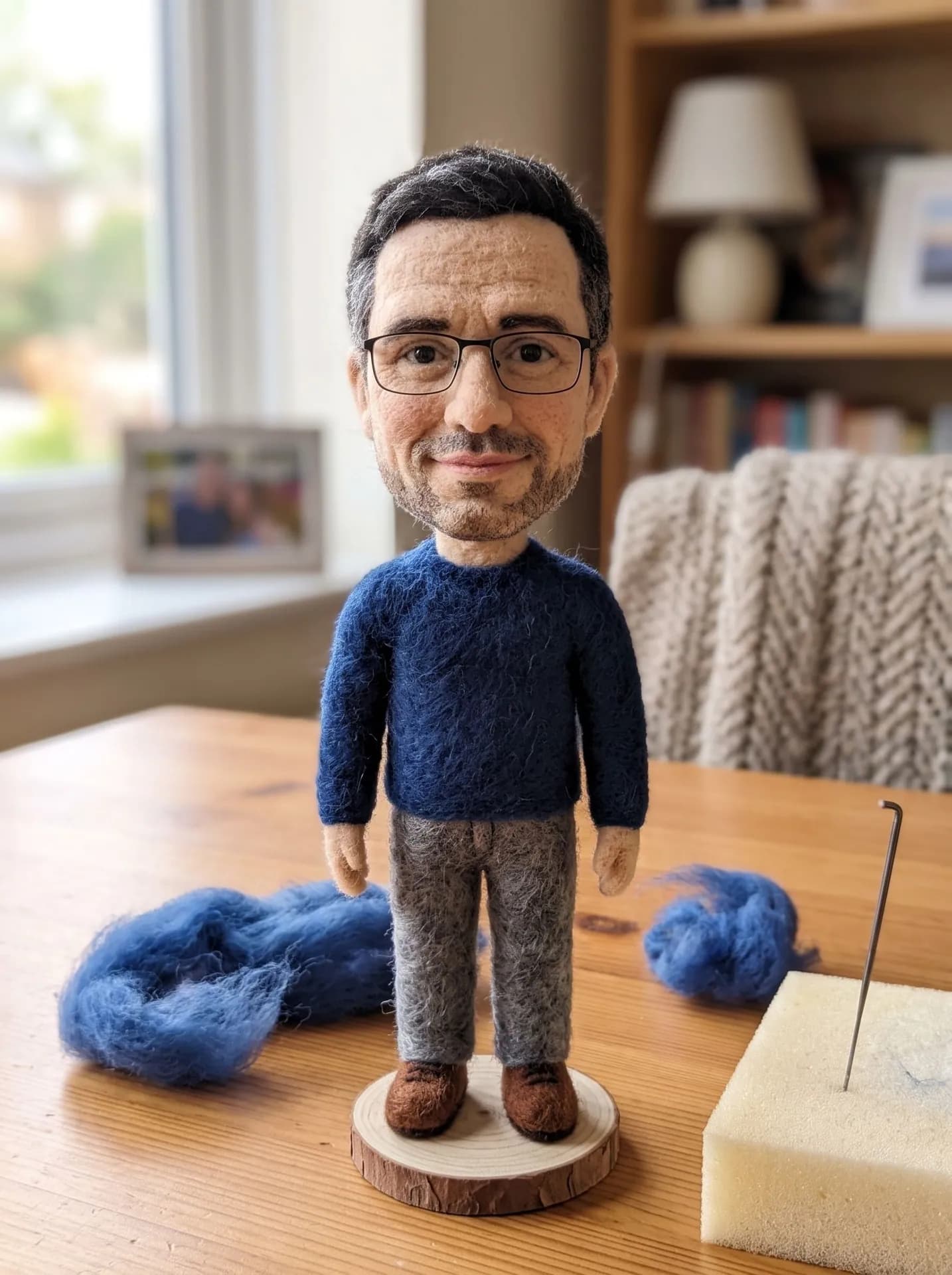 Needle-felted wool figure of a man with glasses and stubble in a blue sweater, standing on a wood slice by a felting needle