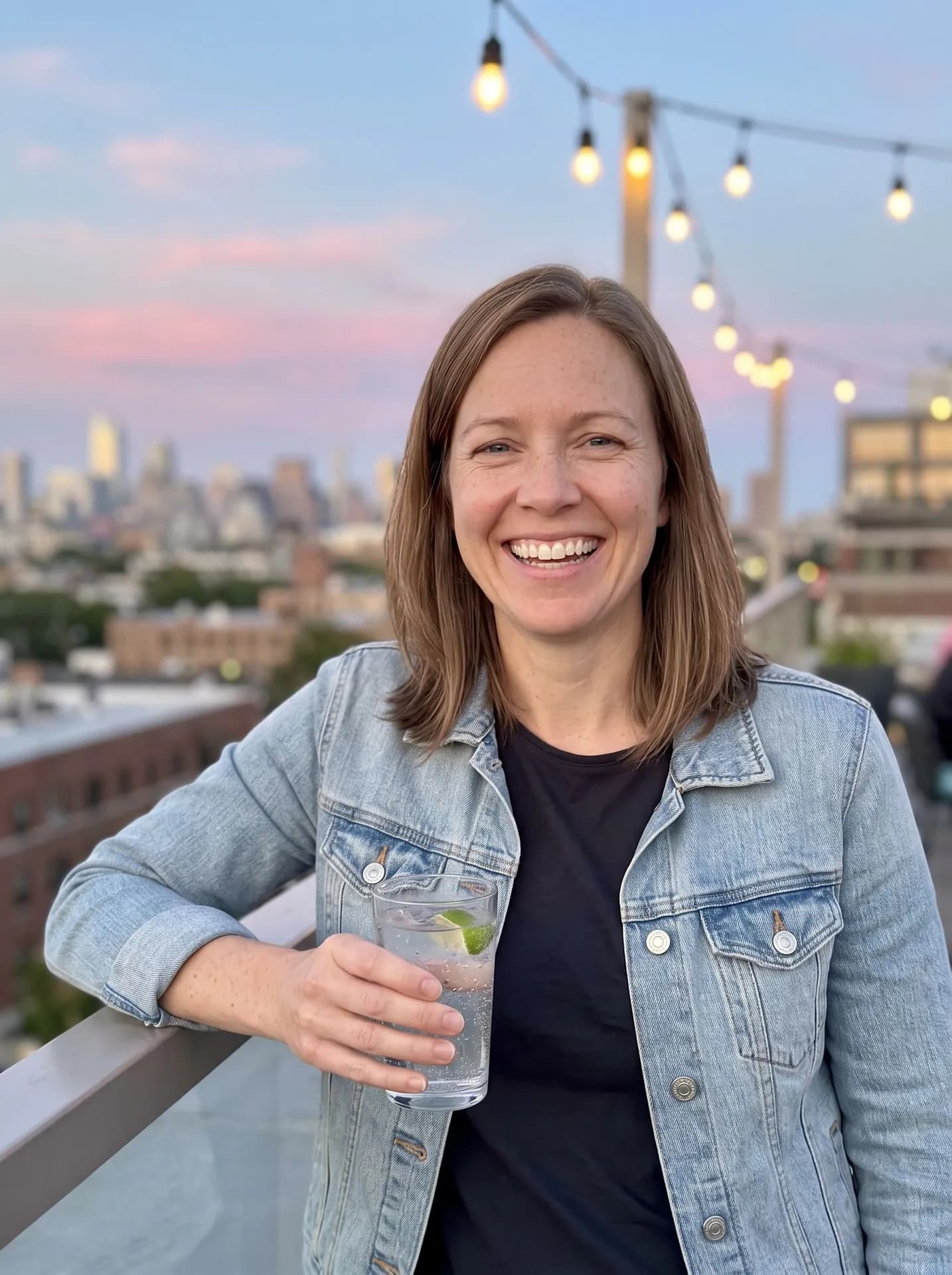 Smiling woman in a denim jacket on a rooftop at dusk, holding a glass of sparkling water with lime under string lights