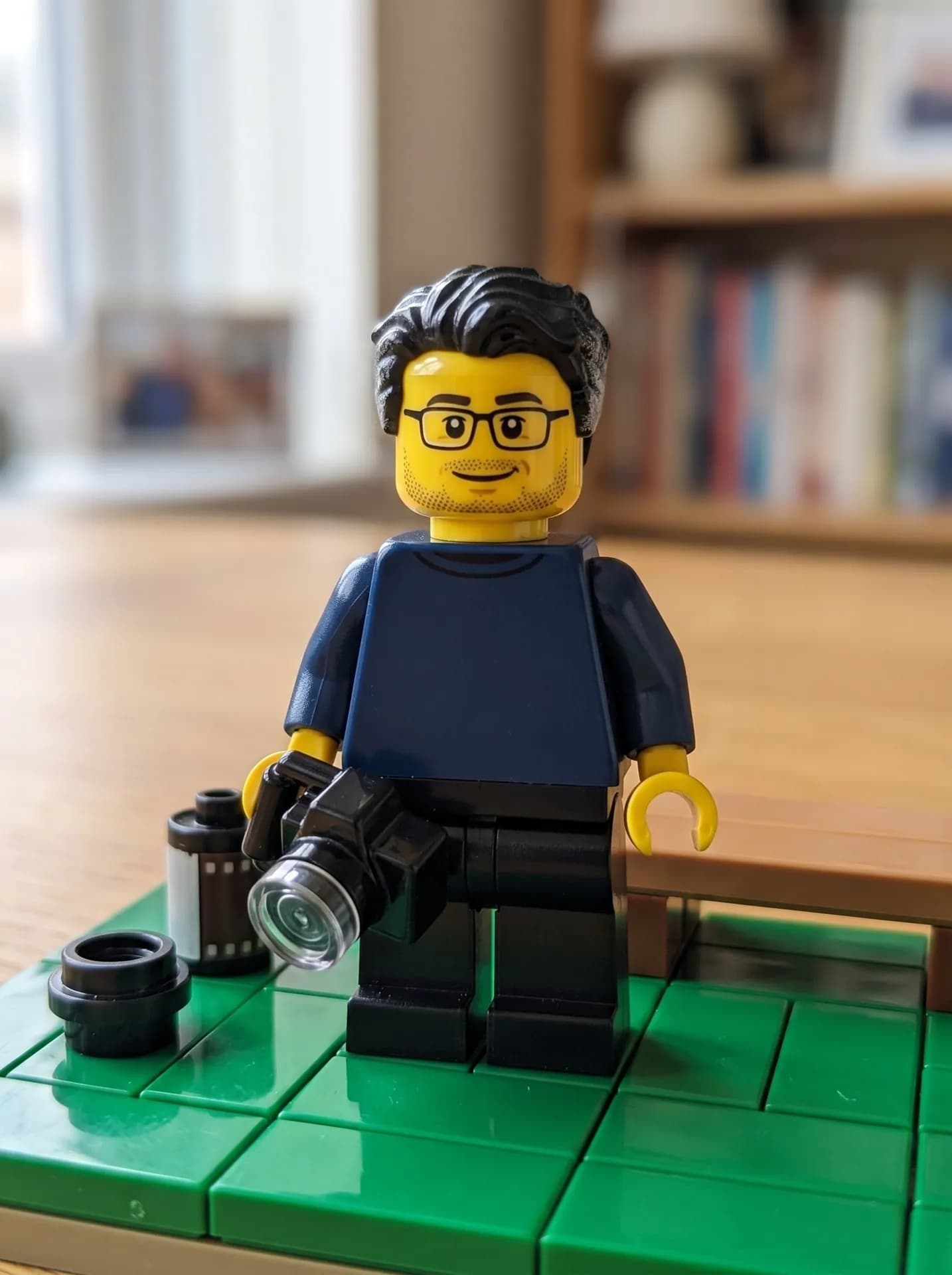 Yellow-head toy minifigure with black hair, printed glasses and stubble in a navy top, holding a tiny camera on green tiles