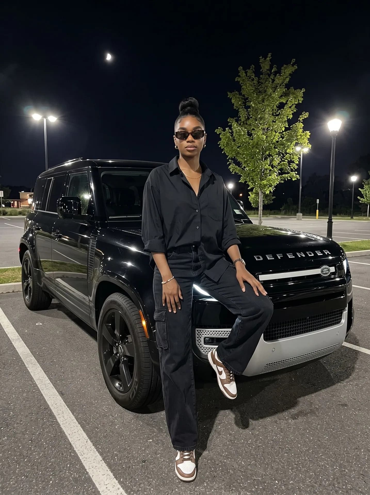 Woman in an oversized black shirt and cargo jeans on the fender of a black Defender at night, sunglasses on
