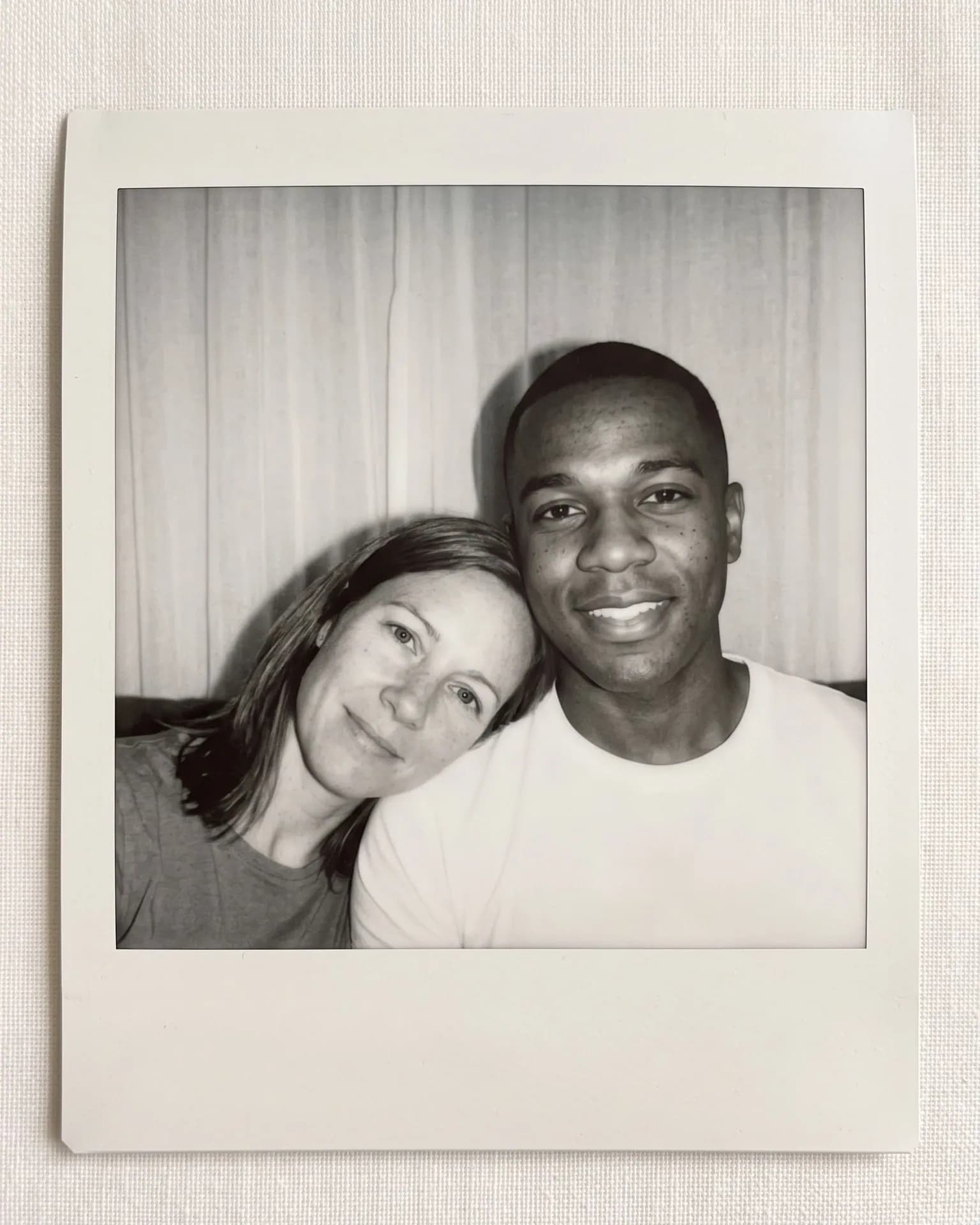 Black-and-white instant print: a woman rests her head on a smiling man's shoulder in front of a white curtain