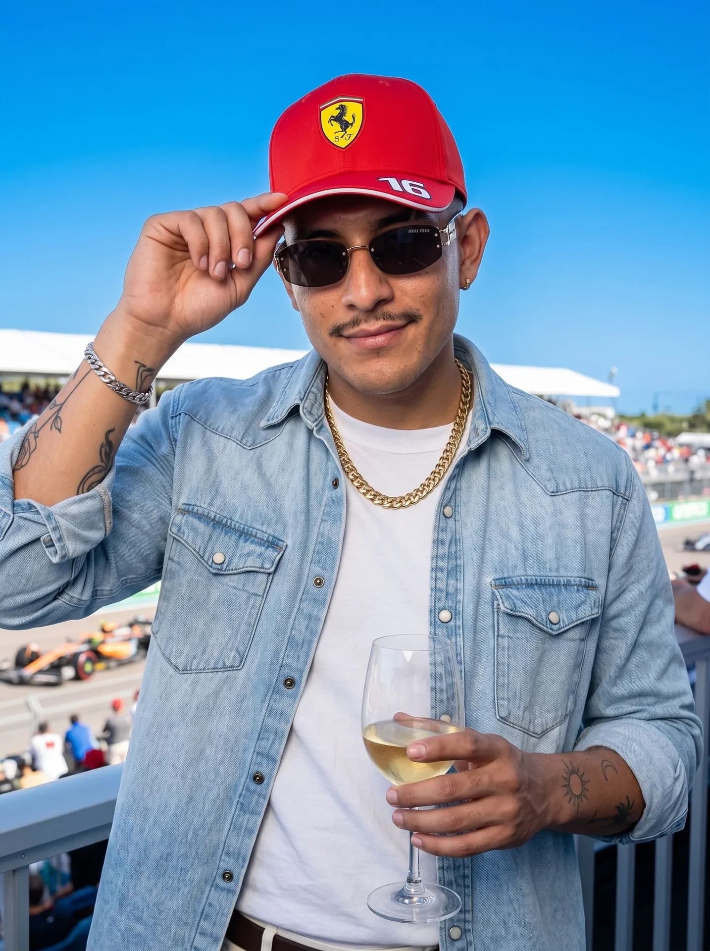 Man with a thin mustache in a red Ferrari cap and denim shirt holding white wine above a racetrack