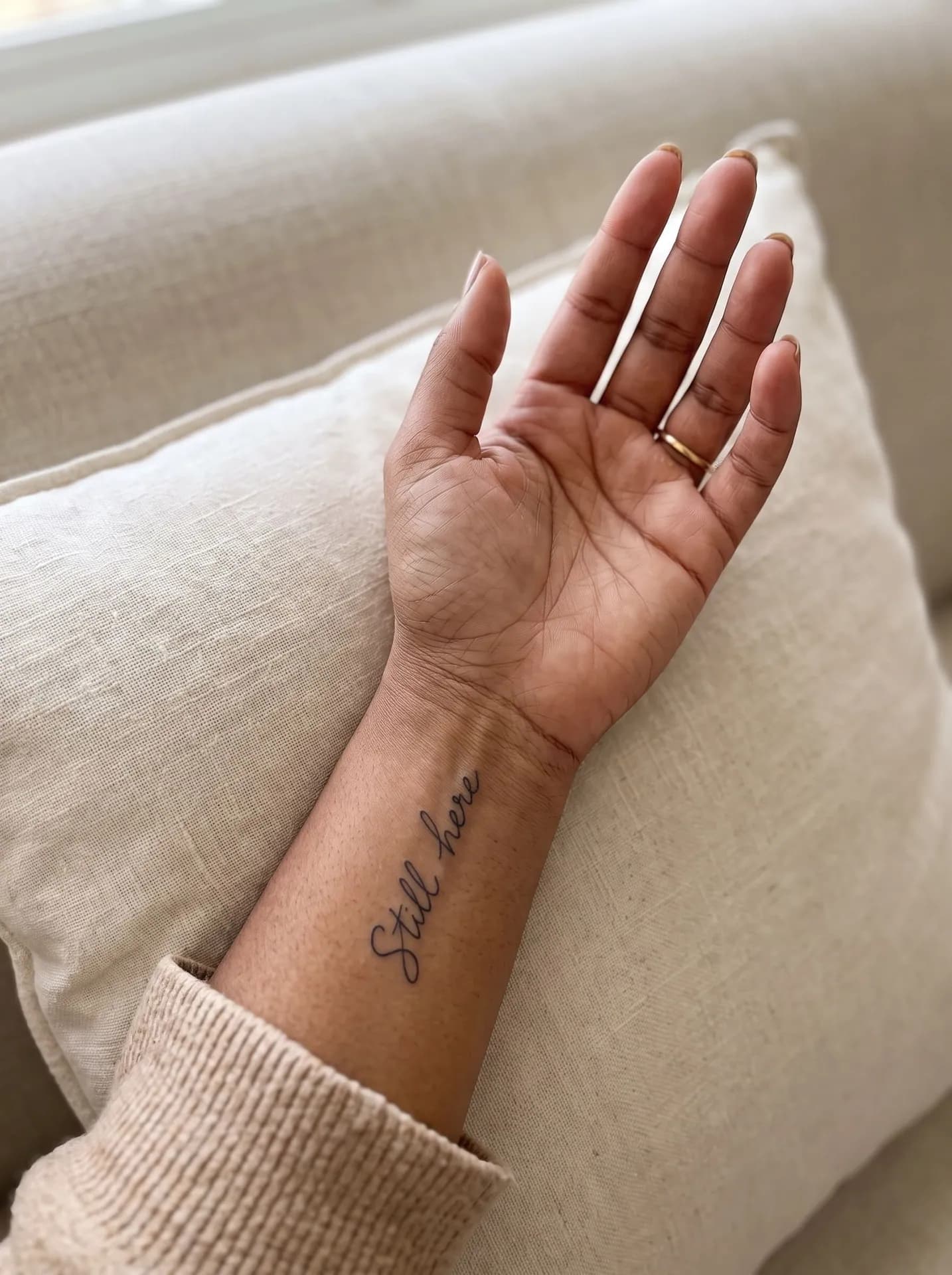 Woman's inner wrist on a linen cushion with a fine cursive tattoo reading Still here along the forearm