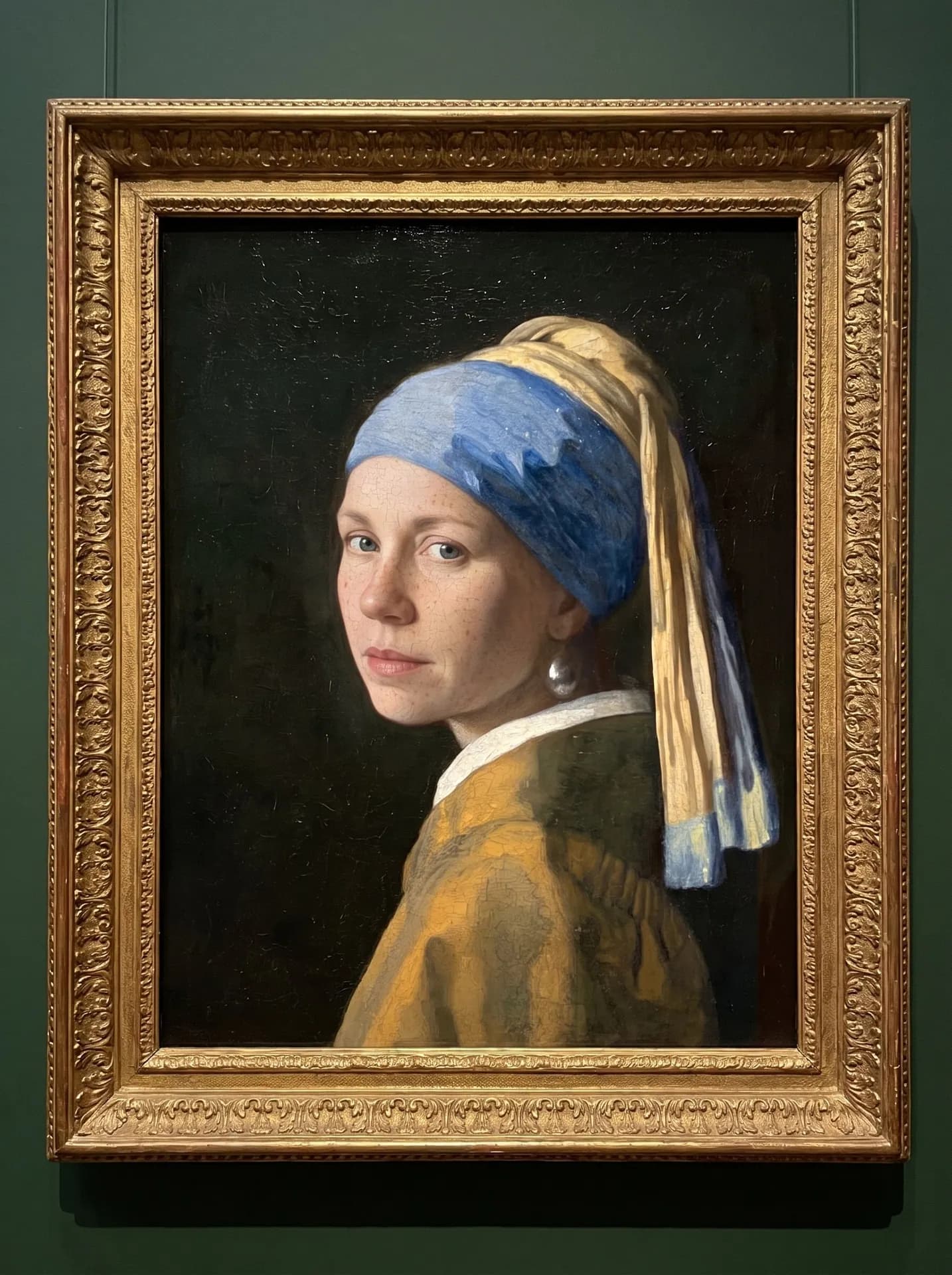 Girl with a Pearl Earring repainted with a blue-eyed, freckled woman as the sitter, in an ornate gilded frame on a green wall