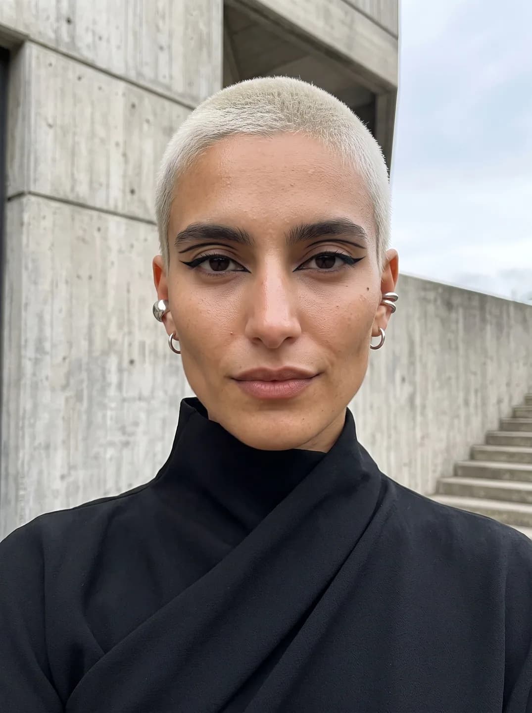 AI-generated sample input: front-facing selfie of a woman with a bleached platinum buzz cut, winged eyeliner and silver ear cuffs in a black high-neck top by a concrete wall