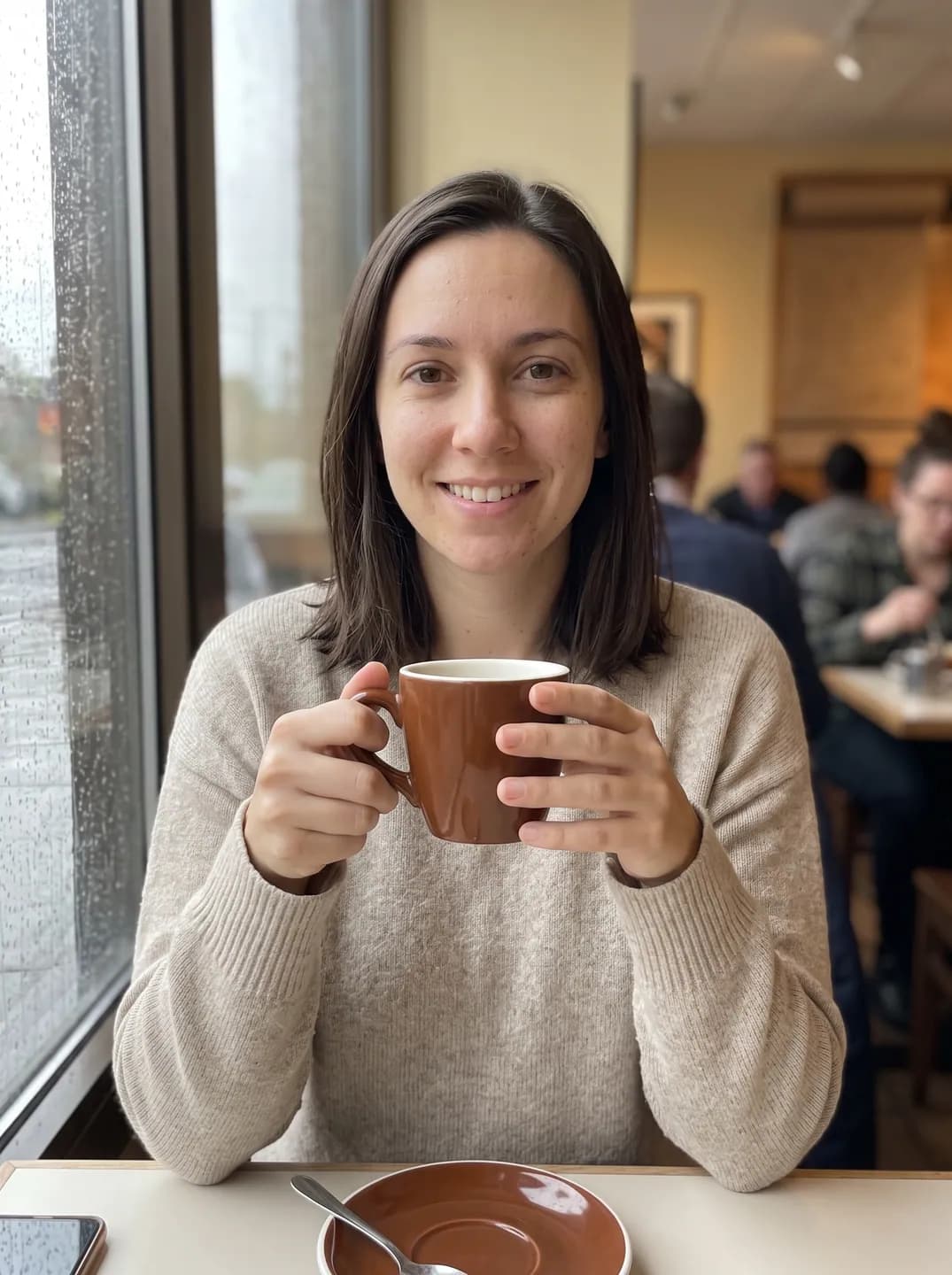 AI-generated sample input: woman in an oatmeal sweater holding a coffee cup at a cafe table by a rainy window