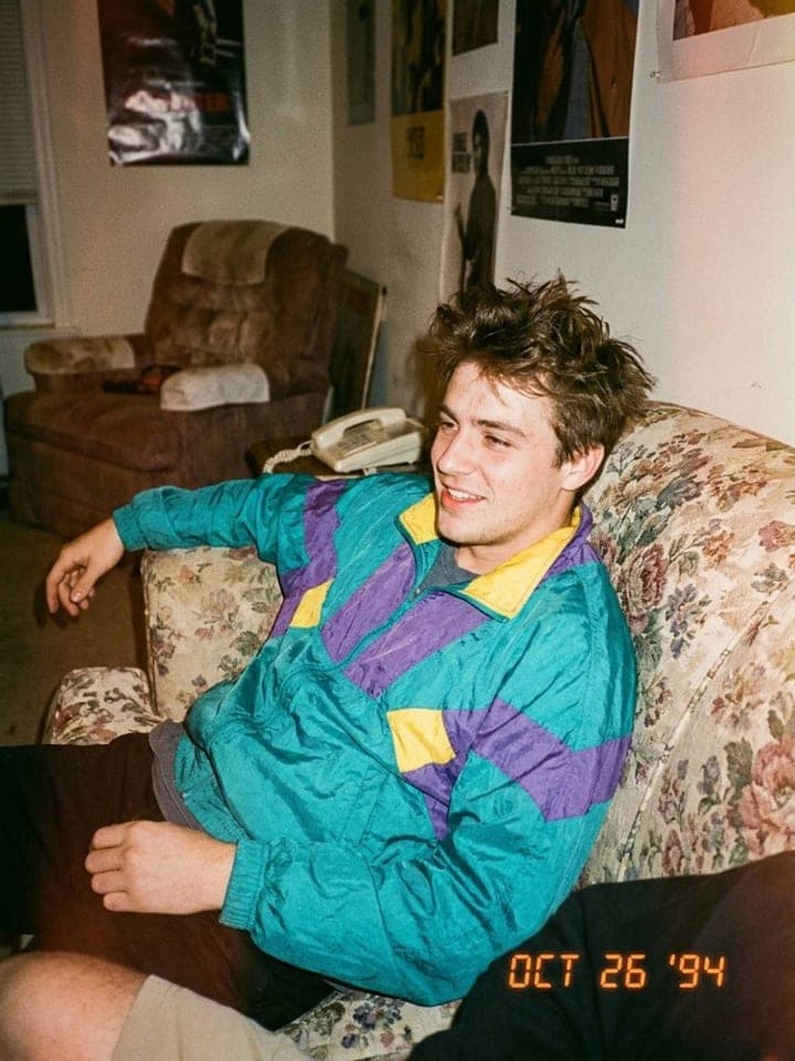 Young man in a bright 90s windbreaker lounging on a floral sofa, orange date stamp OCT 26 '94