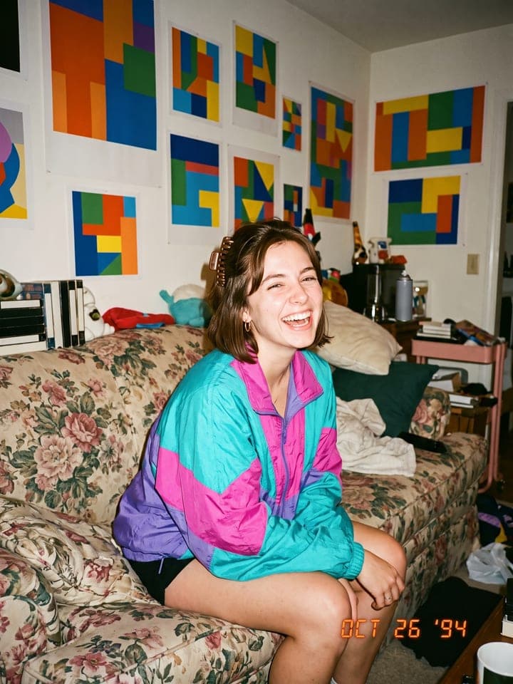 Junge Frau in knalliger 90er-Windjacke lacht auf einem geblümten Sofa, orangefarbener Datumsstempel OCT 26 '94
