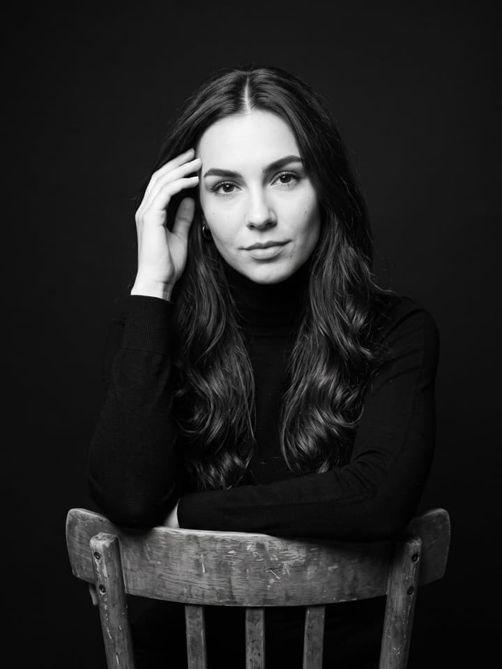 Retrato de estudio en blanco y negro de una mujer de pelo largo, oscuro y ondulado con suéter negro de cuello alto, sentada a horcajadas en una silla de madera, con una mano en la sien, sobre un fondo negro liso