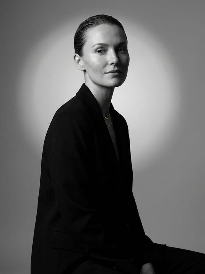 Black-and-white studio portrait of a woman with slicked-back hair in a black blazer, a soft light circle behind