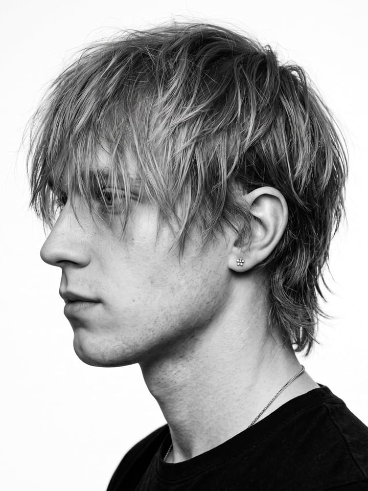Schwarz-Weiß-Seitenprofil eines jungen Mannes mit langen zotteligen blonden Haaren über dem Auge, schwarzes T-Shirt, weißer Hintergrund
