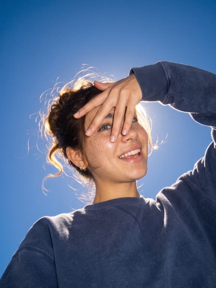 Sommersprossige junge Frau lacht in einem übergroßen marineblauen Fleece-Sweatshirt, von unten vor tiefem kobaltblauem Himmel fotografiert, eine Hand über der Stirn gespreizt, ein Auge gegen die Sonne zugekniffen, lose Strähnen ihres dunklen Messy Buns leuchten im Gegenlicht