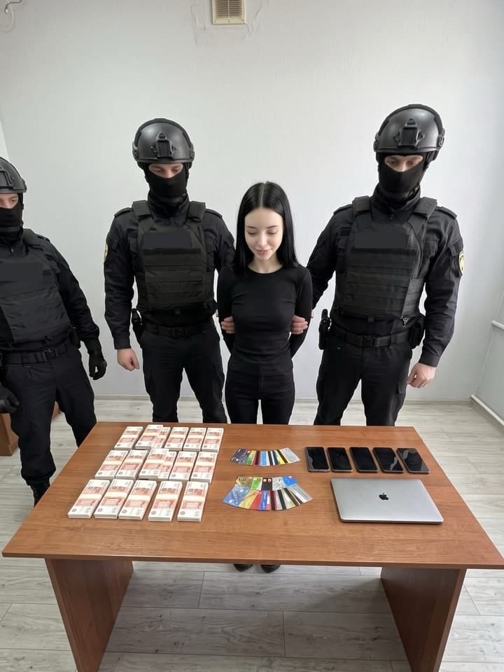 Foto de redada en broma: una mujer de pelo oscuro, vestida de negro y sonriente, sujeta por agentes encapuchados con equipo negro, detrás de una mesa con fajos de billetes, tarjetas y teléfonos