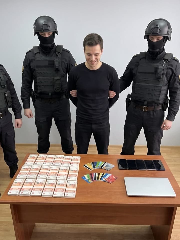 Foto de redada en broma: un hombre joven sonriente vestido de negro, sujeto por dos agentes encapuchados, detrás de una mesa con fajos de billetes, tarjetas bancarias, teléfonos y una laptop