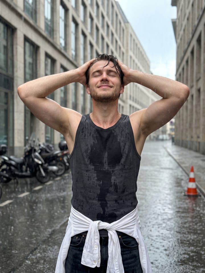 Hombre joven empapado, con los ojos cerrados, echándose el pelo mojado hacia atrás con las dos manos en una calle de la ciudad bajo la lluvia