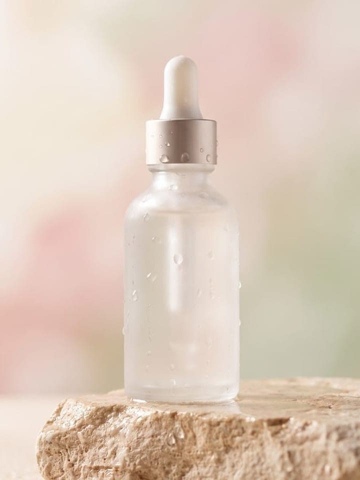 Frosted serum bottle with a dropper on a stone podium with water droplets and a pastel background