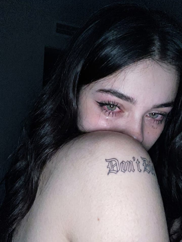 Dark close-up of a woman with teary eyes, her shoulder with a Don't Die tattoo covering her lower face