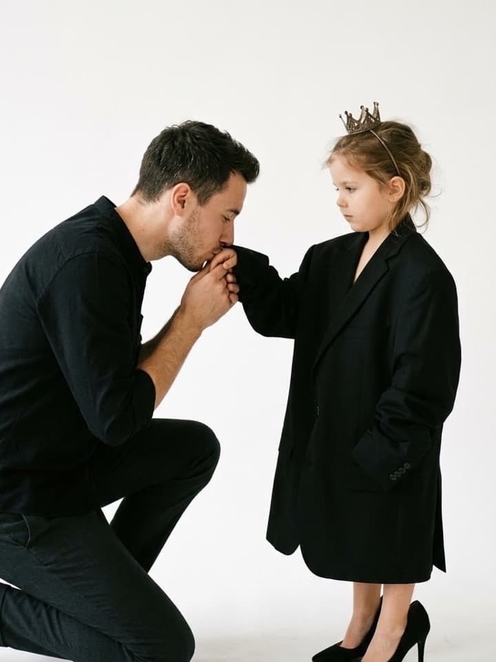 Foto de estudio de un hombre arrodillado besando la mano de una niña con un blazer negro extragrande, tacones negros y una pequeña corona