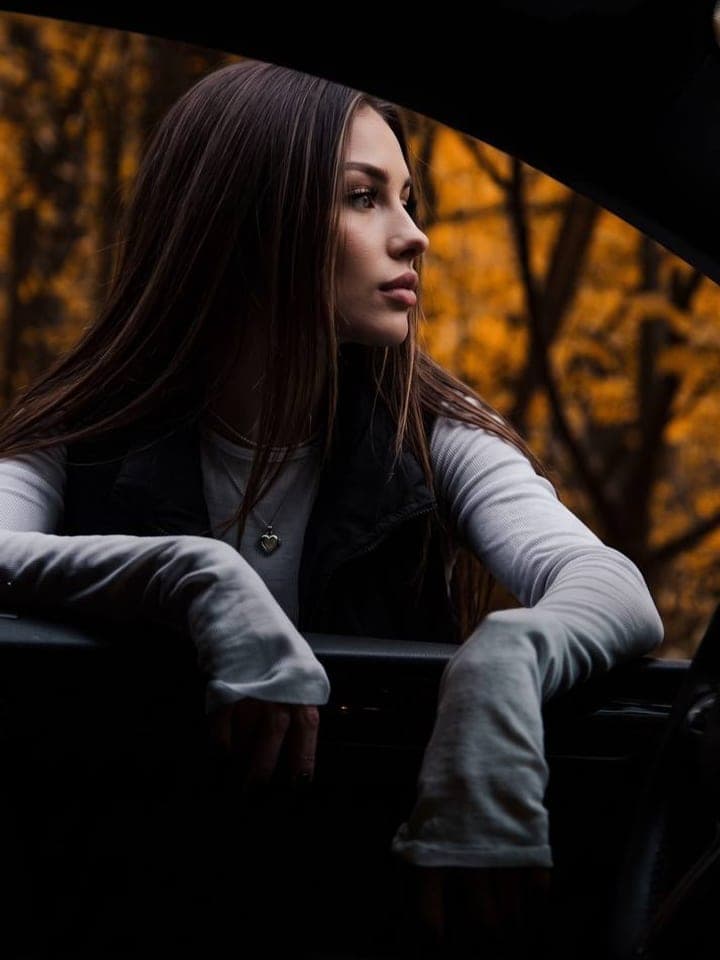 Woman with long dark hair leaning out of a dark car window, golden autumn forest behind her