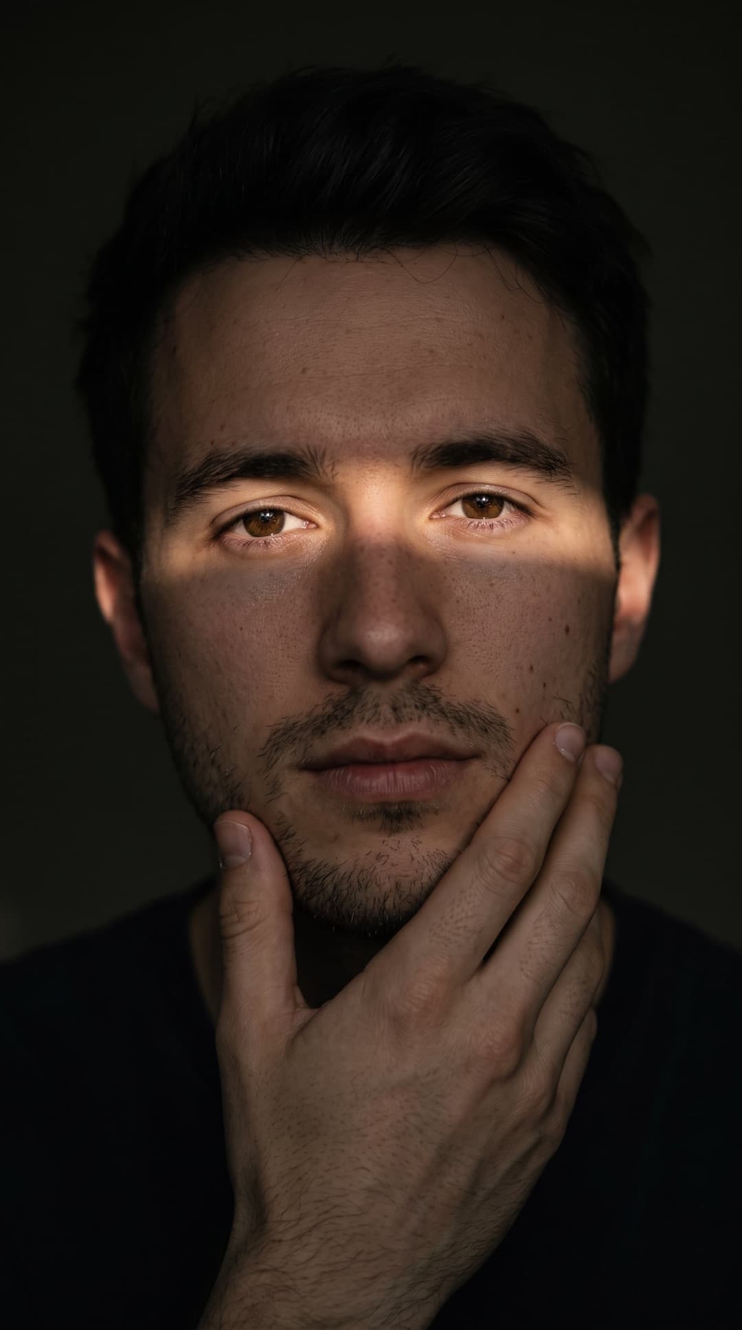 Close de um jovem de cabelo escuro num quarto escuro, com uma faixa de luz quente sobre os olhos e a mão no queixo