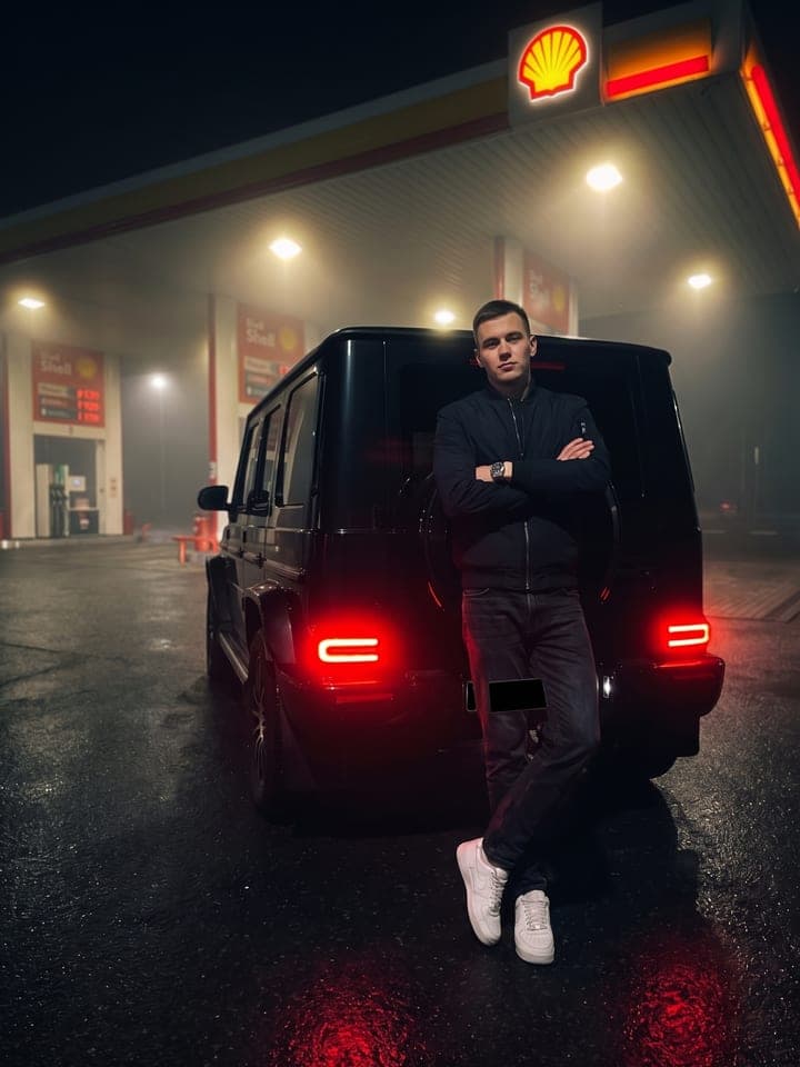 Young man in a bomber leaning on a black Mercedes G-Class at a foggy Shell gas station at night