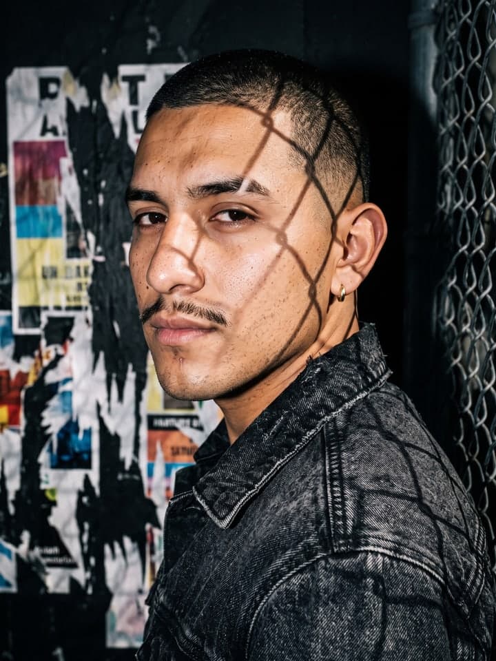 Hombre con el pelo rapado y un bigote fino, con chaqueta denim en negro lavado, mirando hacia atrás por encima del hombro bajo un flash duro, con la sombra en rombos de una valla de alambre sobre un lado de la cara y carteles de colores rotos en una pared oscura detrás