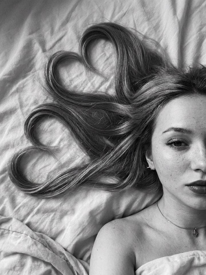Black-and-white photo of a woman lying on sheets, her long hair arranged into heart shapes