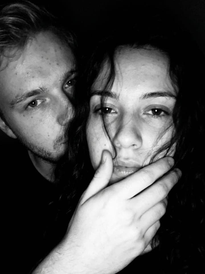 Dark black-and-white photo of a couple, his hand softly covering her lips as both look into the camera