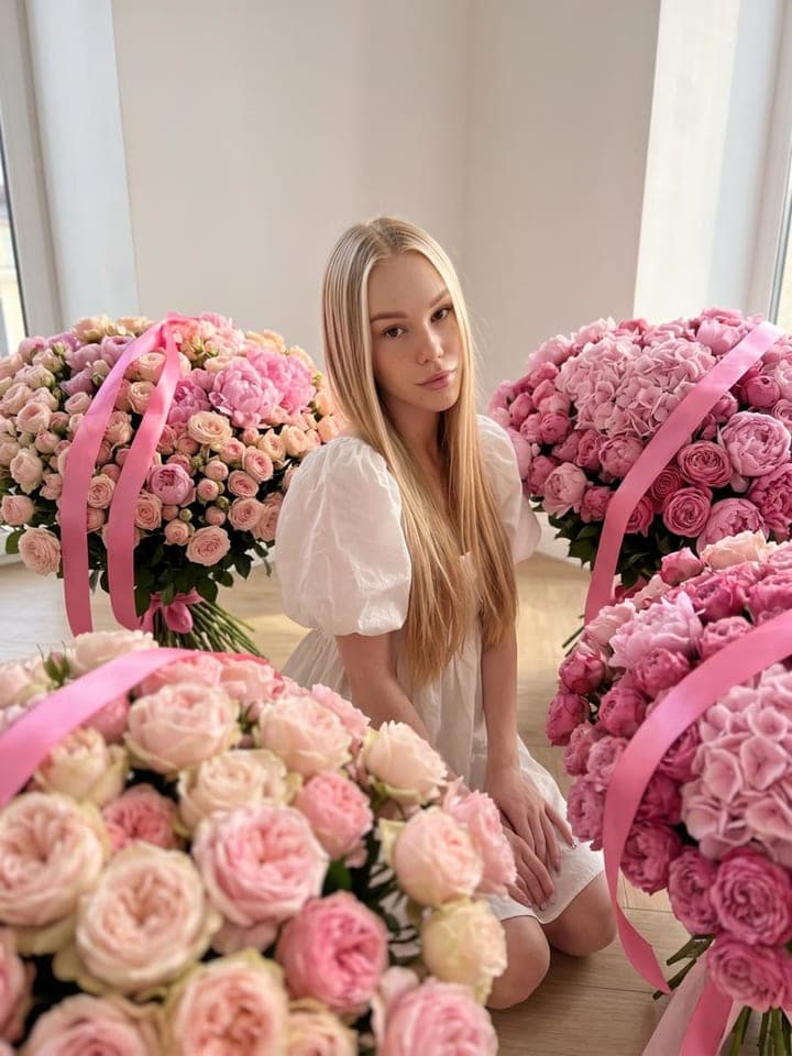 Blonde woman in a white dress kneeling among huge bouquets of pink roses with pink satin ribbons