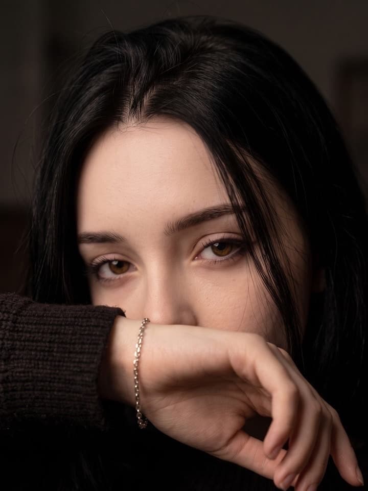 Nahaufnahme einer dunkelhaarigen Frau, deren Hand und silbernes Gliederarmband Nase und Mund verdecken, nur ihre Augen sind zu sehen, dunkler Hintergrund