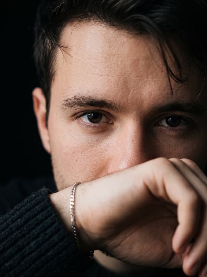 Nahaufnahme eines jungen Mannes mit dunklen Haaren, dessen Hand und silbernes Armband Nase und Mund verdecken, nur seine Augen sind zu sehen