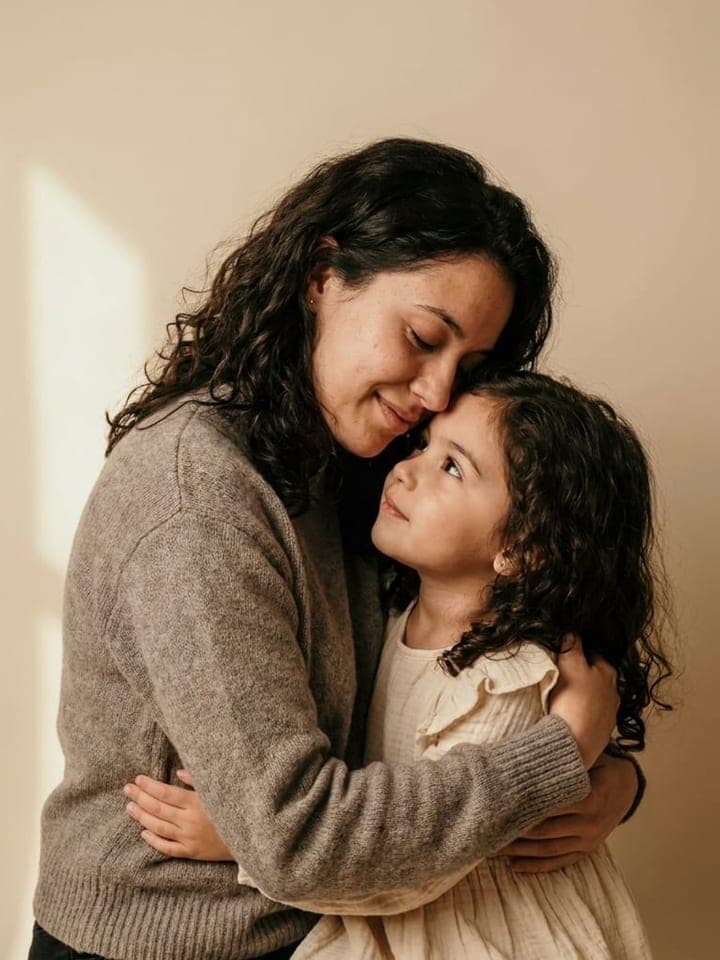 Woman with dark curly hair hugging a little girl with the same curls against a warm beige backdrop