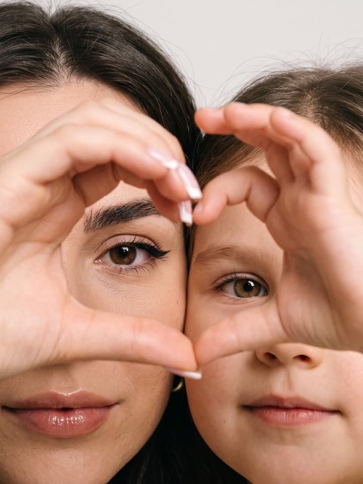 Nahaufnahme einer Mutter und ihrer kleinen Tochter Wange an Wange, von jedem Gesicht die Hälfte im Bild, ihre Hände formen ein Herz um ihre Augen