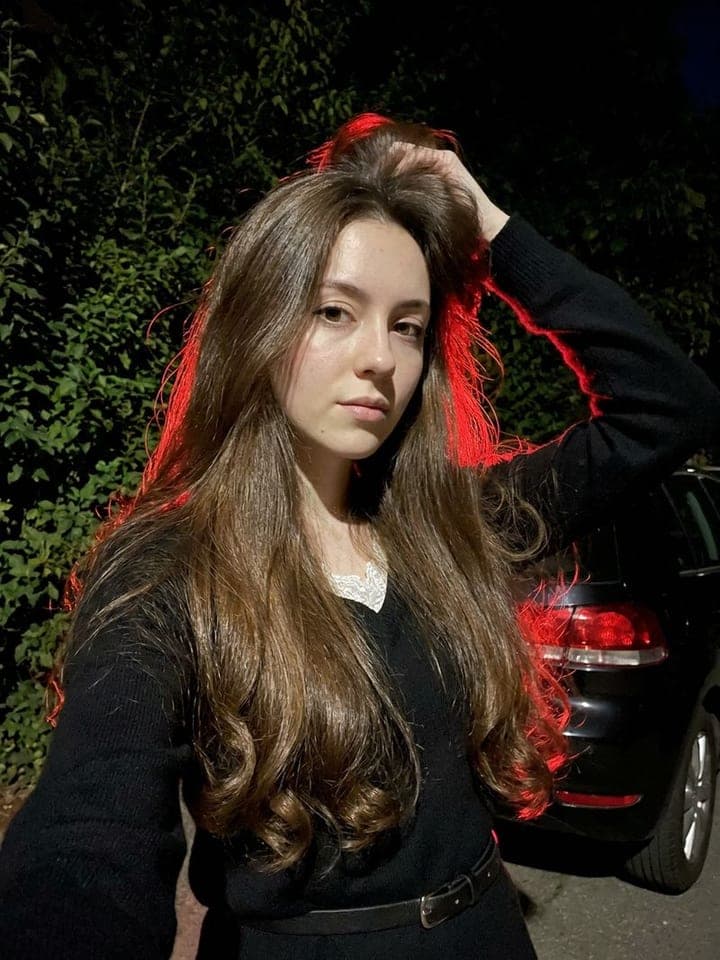 Woman with long brown hair lifting it with one hand, red car light glowing behind her at night