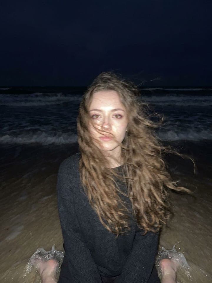 Woman with long windblown hair sitting on dark sand at night, lit by phone flash against a black sea