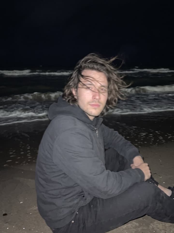Hombre joven con el pelo largo al viento y una sudadera oscura con capucha, sentado en la arena oscura de noche, iluminado por el flash del teléfono frente a un mar negro