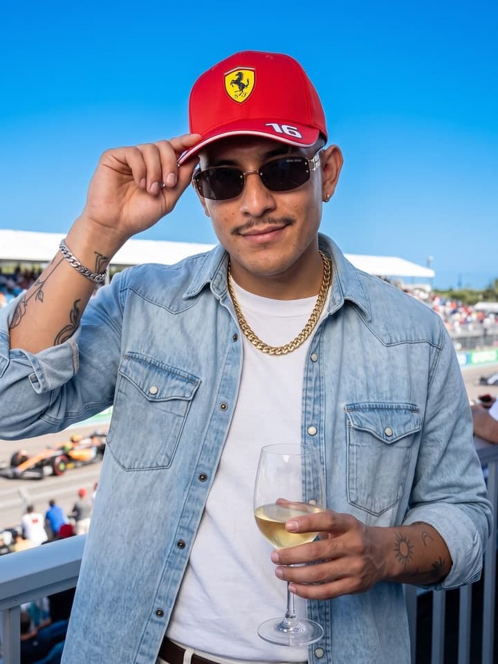Hombre con bigote fino en una tribuna VIP de un circuito soleado, ajustándose una gorra roja de Ferrari con un 16 blanco en la visera, con gafas de sol oscuras y una camisa denim clara sobre una camiseta blanca con una cadena de oro, sosteniendo una copa de vino blanco, con autos de carreras en la pista detrás