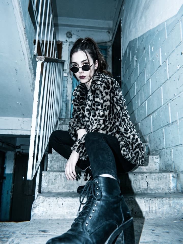 Blitzfoto von unten einer jungen Frau mit runder Sonnenbrille und Kunstpelzmantel im Leopardenmuster, die auf einer Betontreppe hockt, vorn ein Stiefel