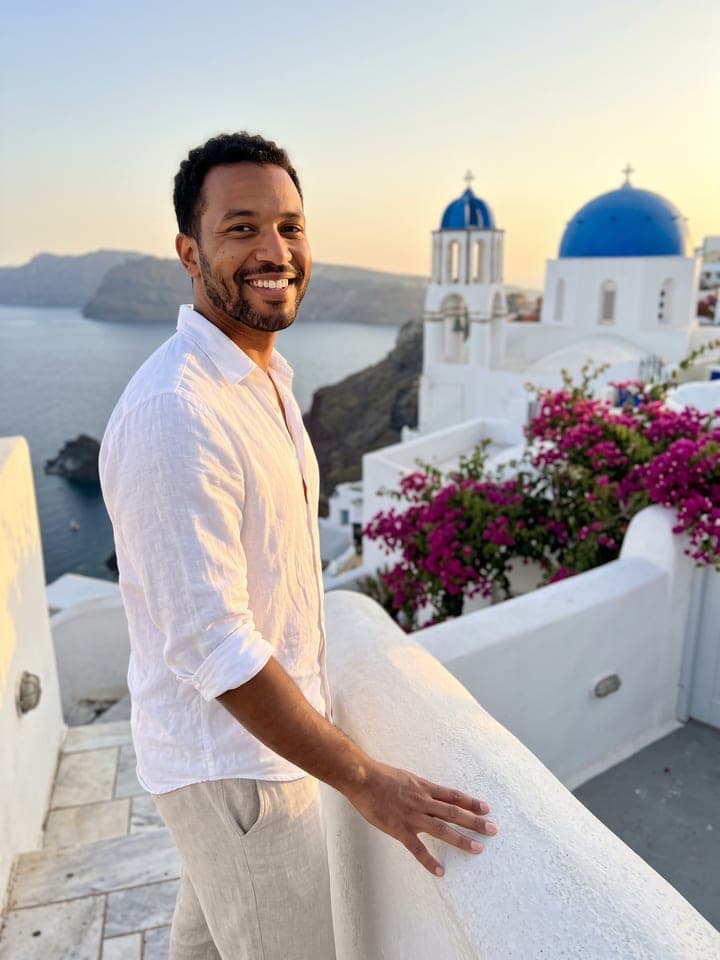 Hombre sonriente de pelo negro, corto y rizado, con la barba arreglada y una camisa blanca de lino, en una terraza encalada de Santorini en la hora dorada, con una iglesia de cúpula azul, buganvillas y el mar de la caldera a su lado