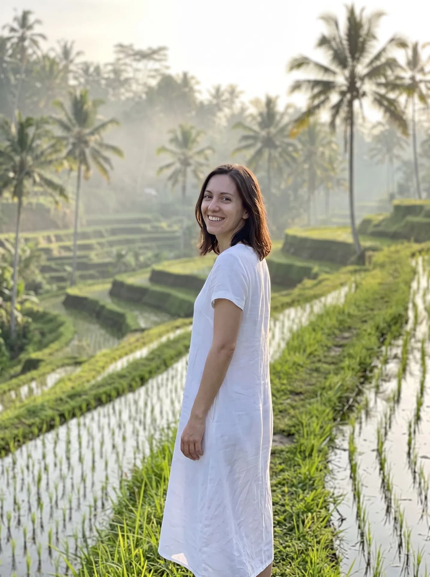 Smiling woman in a white linen dress glancing back on a grassy ridge between misty rice terraces and palm trees