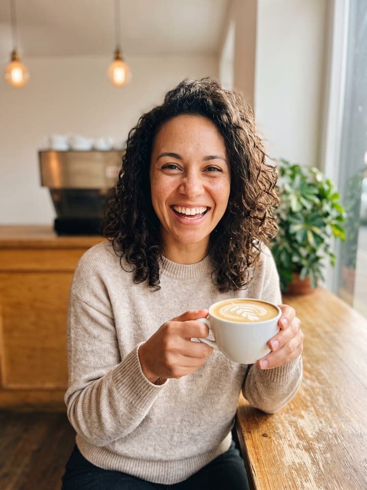 Spontanes Handyfoto einer lachenden Frau Anfang dreißig mit schulterlangen dunklen Locken und Sommersprossen im haferfarbenen Strickpulli, die in einem sonnigen Café an einem Holztisch einen Latte hält