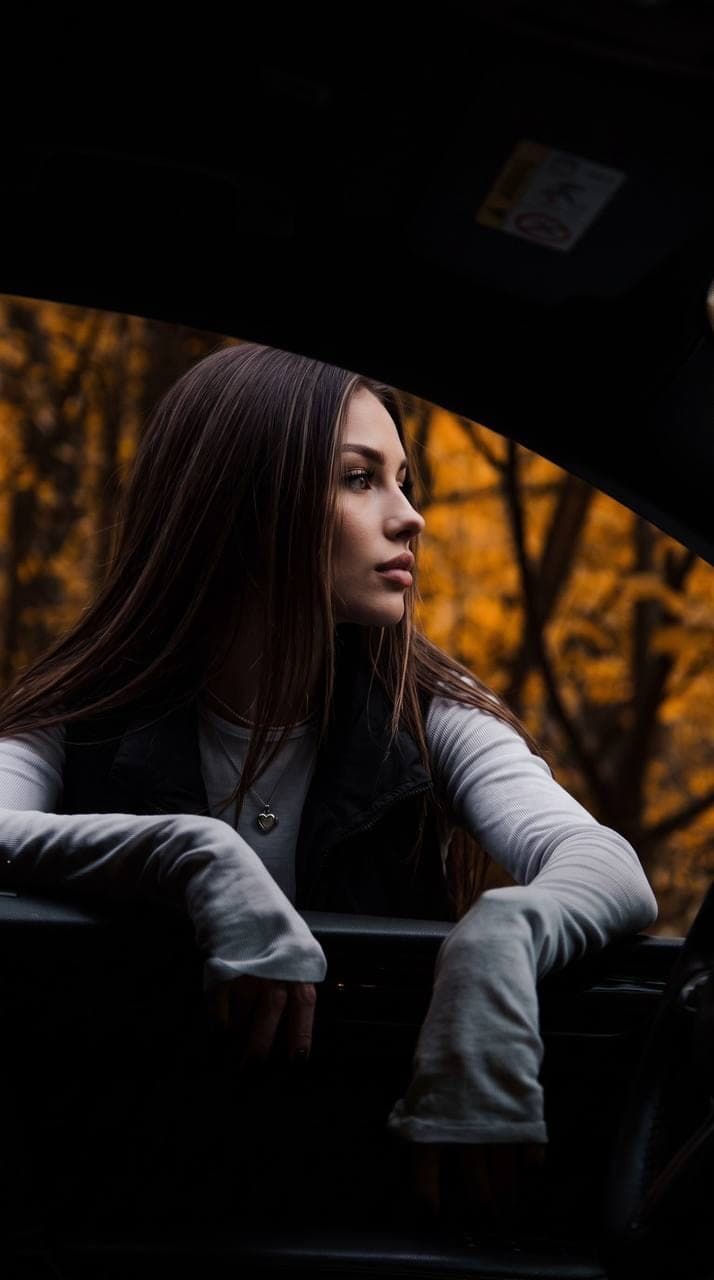 Frau mit langen dunklen Haaren lehnt sich aus einem dunklen Autofenster, hinter ihr ein goldener Herbstwald