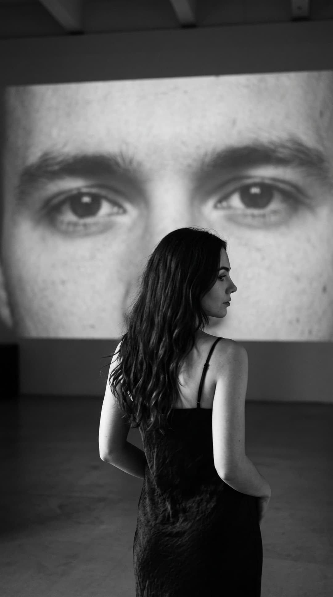 Schwarz-Weiß-Foto einer dunkelhaarigen Frau im schwarzen Slipkleid, mit dem Rücken zur Kamera, vor einer riesigen Projektion der Augen eines Mannes