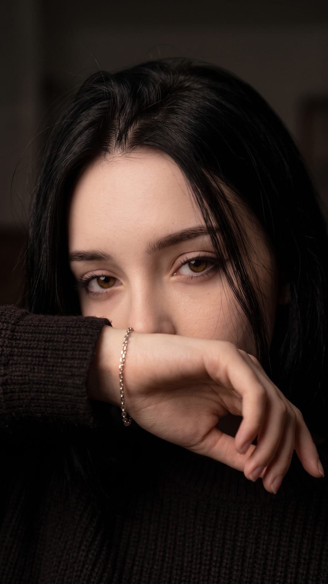 Nahaufnahme einer dunkelhaarigen Frau, deren Hand und silbernes Gliederarmband Nase und Mund verdecken, nur ihre Augen sind zu sehen, dunkler Hintergrund