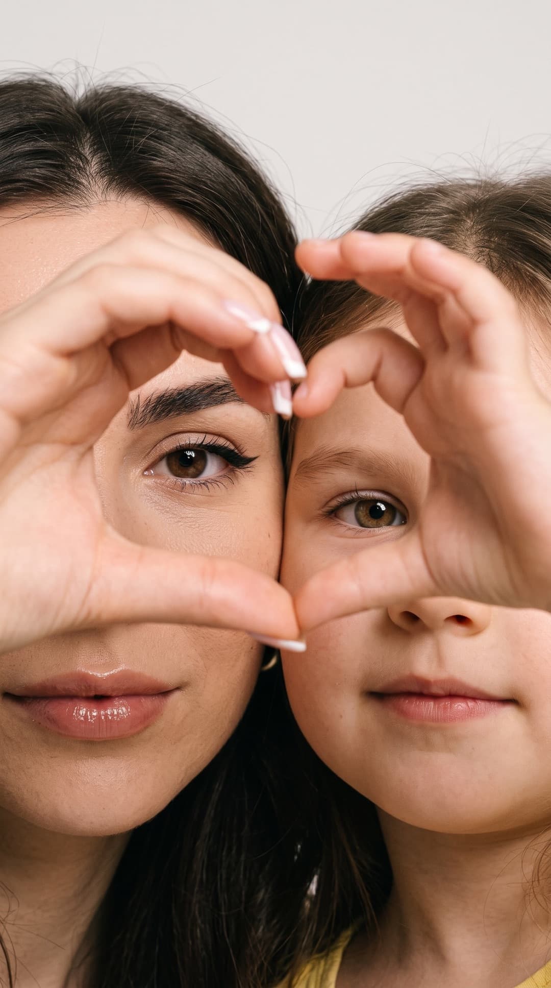 Nahaufnahme einer Mutter und ihrer kleinen Tochter Wange an Wange, von jedem Gesicht die Hälfte im Bild, ihre Hände formen ein Herz um ihre Augen