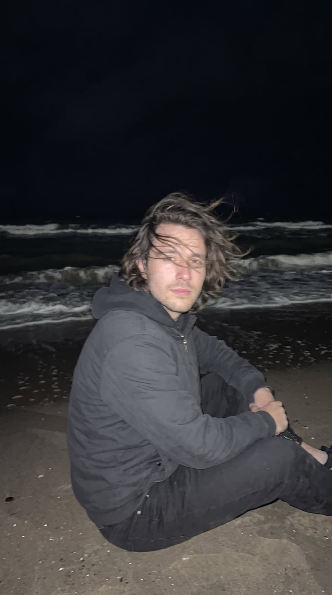 Jovem de cabelo longo ao vento e moletom escuro sentado na areia escura à noite, iluminado pelo flash do celular diante de um mar negro