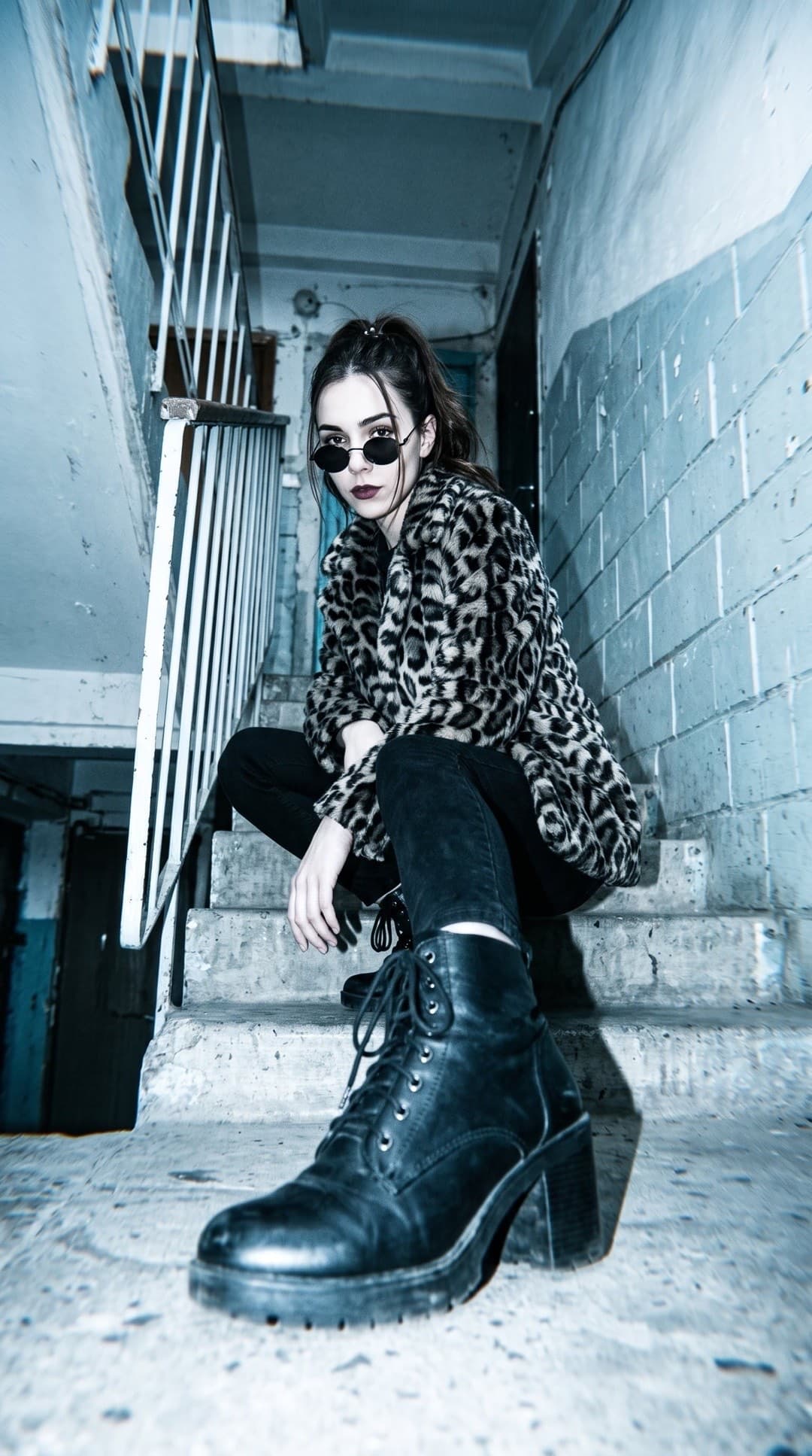 Blitzfoto von unten einer jungen Frau mit runder Sonnenbrille und Kunstpelzmantel im Leopardenmuster, die auf einer Betontreppe hockt, vorn ein Stiefel