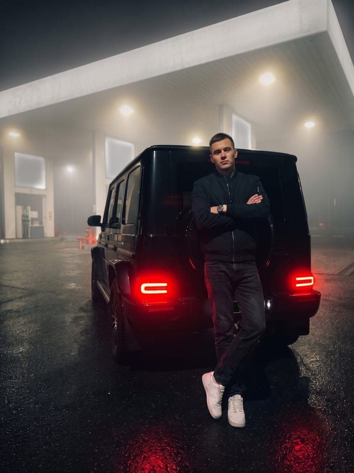 Junger Mann in Bomberjacke lehnt nachts an einer schwarzen Mercedes G-Klasse an einer nebligen Tankstelle