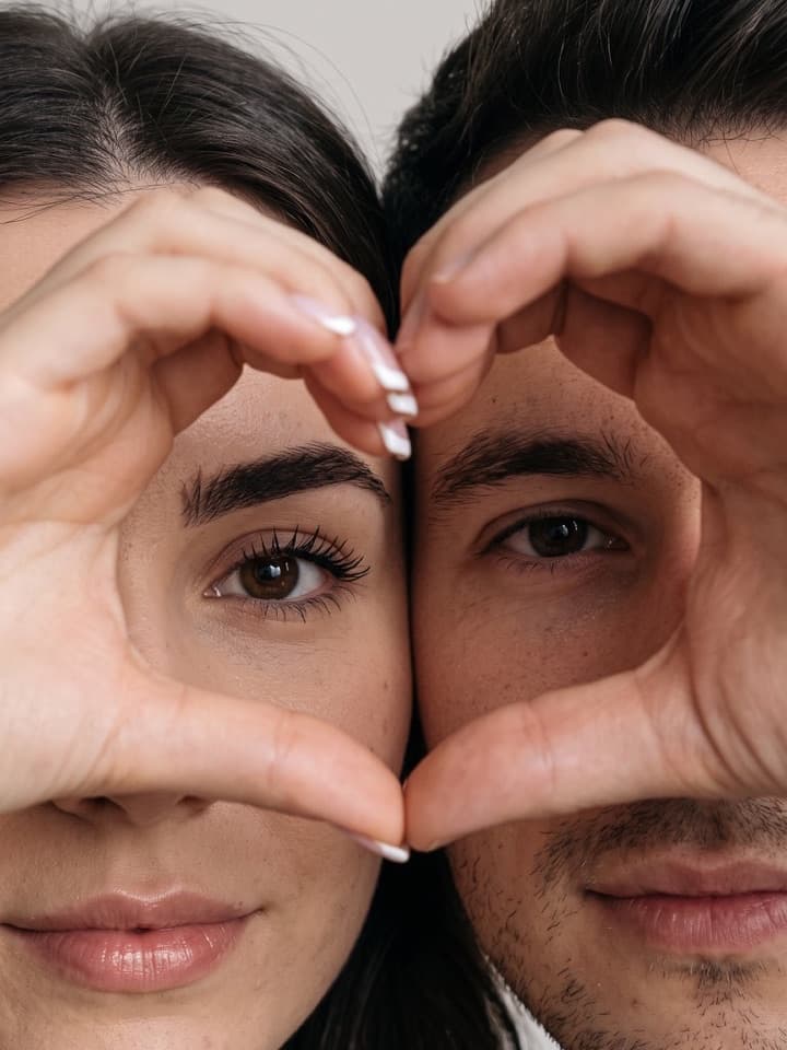 Nahaufnahme einer Frau und eines Mannes Wange an Wange, von jedem Gesicht die Hälfte im Bild, ihre Hände formen ein Herz um ihre Augen